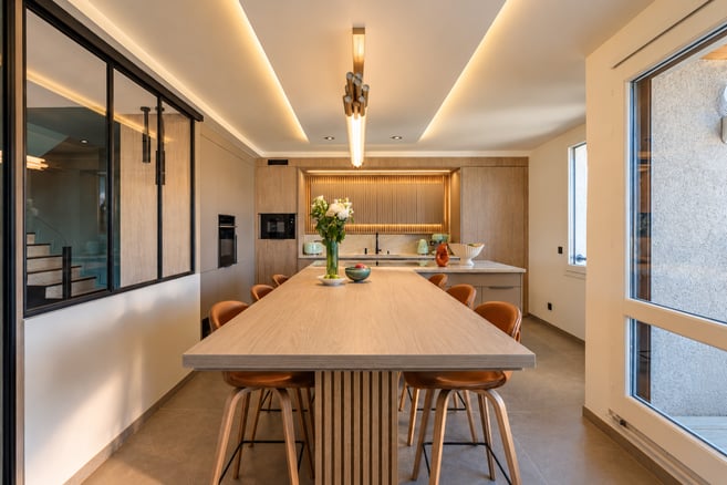 Cuisine contemporaine aux tons naturels, associant bois clair, mobilier épuré et verrière noire. Une grande table centrale, entourée de chaises en cuir cognac, structure l’espace, sublimé par des éclairages indirects chaleureux.