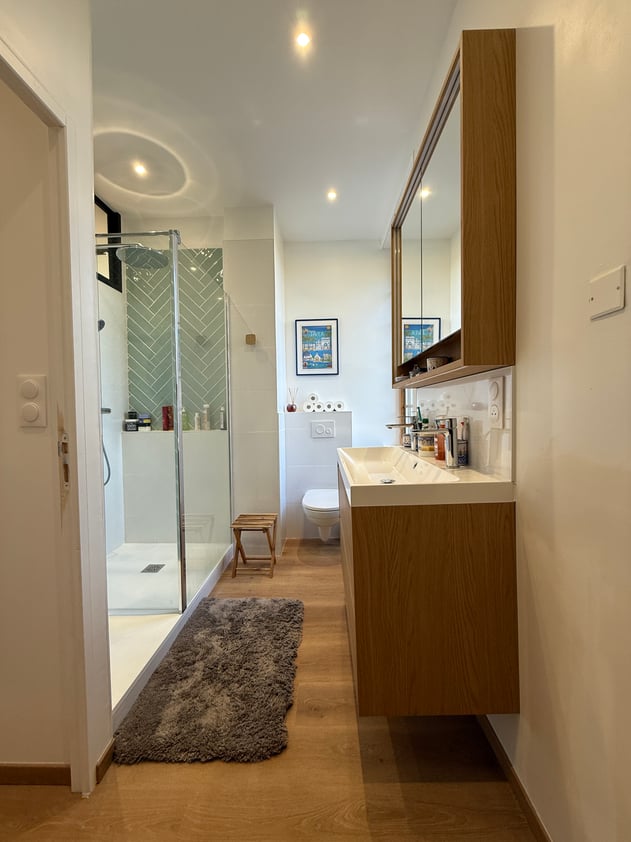La salle de bain parentale, pensée dans la longueur — douche italienne en chevron vert sauge, meuble chêne miel, fonctionnelle et élégante.