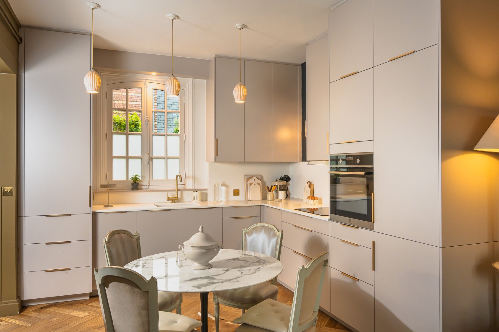 Vue d’ensemble de la cuisine sur mesure aux façades claires, poignées dorées et table ronde, réalisée par PERENE Caen à Villers-sur-Mer. Vue d’ensemble de la cuisine sur mesure aux façades claires, poignées dorées et table ronde, réalisée par PERENE Caen à Villers-sur-Mer.