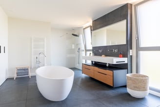 Dans la suite parentale, une grande salle-de-bain permet de prendre ses aises. La baignoire, ovale, occupe le devant de la scène et fait face à une grande baie ouvrant sur le jardin. Un meuble, suspendu pour plus de légèreté, en bois,  est élégamment entouré de noir. les vasques sont elles aussi suspendues et surplombées d'un large miroir.