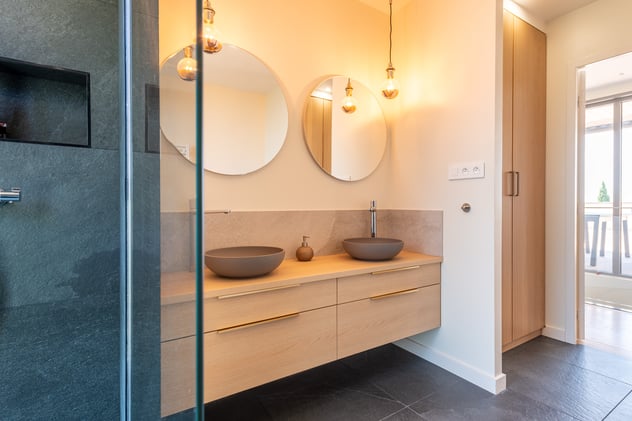 Dans la suite parentale, une grande salle-de-bain permet de prendre ses aises. La baignoire, ovale, occupe le devant de la scène et fait face à une grande baie ouvrant sur le jardin. Un meuble, suspendu pour plus de légèreté, en bois,  est élégamment entouré de noir. les vasques sont elles aussi suspendues et surplombées d'un large miroir.